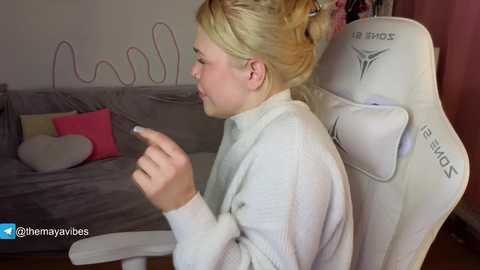 Video of a blonde woman in a white sweater, sitting in a white gaming chair, holding a phone. Background shows a gray sofa with a heart pillow and a pink cushion.