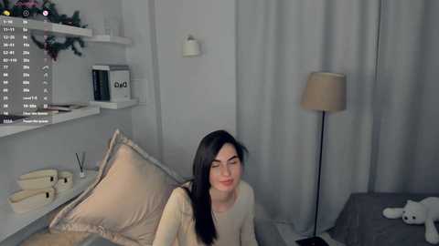 Video of a young woman with long black hair and fair skin, wearing a cream sweater, seated on a beige bed with a white pillow, in a minimalist, modern bedroom with gray curtains and a beige lamp.