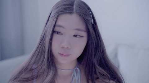 Video of an Asian woman with long, straight, dark brown hair, wearing a light gray halter top, seated in a soft-focus, minimalist setting.