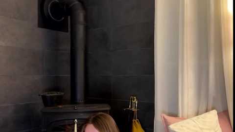 Video of a woman with blonde hair, wearing a yellow blouse, sitting in a dimly lit room with a black metal stove, dark grey tiled wall, and white curtain.