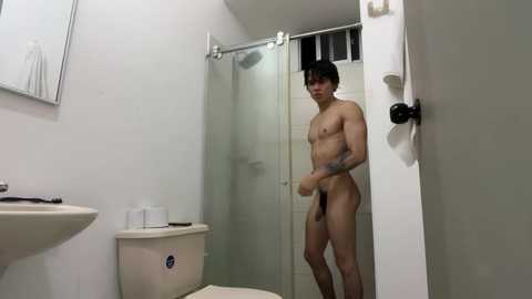 Video of a young, naked Asian man with short black hair standing in a small, white-tiled bathroom. He has a lean build and small, erect penis. The bathroom includes a white toilet, sink, and shower.