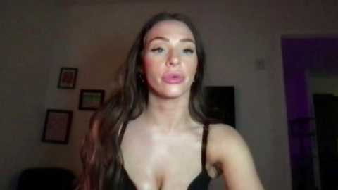 A video of a fair-skinned woman with long, wavy brown hair, wearing a black lace bra, puckering her lips in a seductive pose. The background features dimly lit walls with framed pictures and a partially visible doorway.