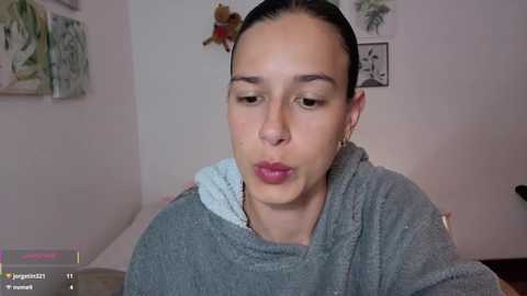 Video of a light-skinned woman with straight black hair, wearing a grey hoodie, making a kissy face indoors. Background includes green and black framed art, teddy bear, and bed.