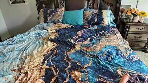 Video of a bed with a turquoise and gold marble-patterned duvet, two matching pillows, and a wooden headboard. Nightstand with a vase of yellow flowers and a lamp beside it.