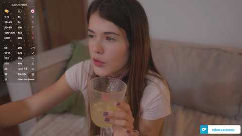 A video of a young woman with long brown hair, wearing a white top, sipping a glass of juice, sitting on a beige couch, background showing a TV screen displaying \"Lounging\" mode.