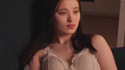 Media: Video of a young woman with fair skin and long, dark hair, wearing a white lace sleeveless top, looking thoughtful against a dark, textured wall.