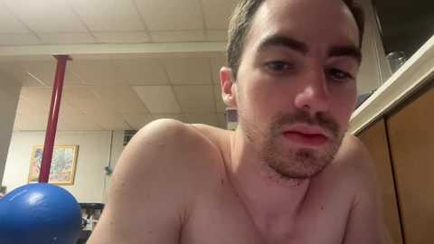 A video of a shirtless man with a beard, light skin, and short brown hair, leaning forward. Background includes a red pole, a blue inflatable object, and a blurred wall with framed art.