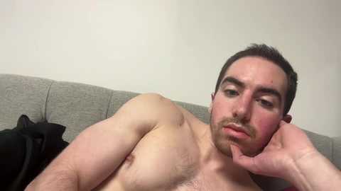 A video of a shirtless, fair-skinned man with short dark hair and a trimmed beard, lying on a grey couch with a black pillow. He has a pensive expression, resting his head on his hand.
