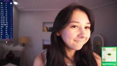 Video of a smiling East Asian woman with long black hair, wearing a sleeveless top, in a cozy, dimly lit room with a TV showing a sports scoreboard.