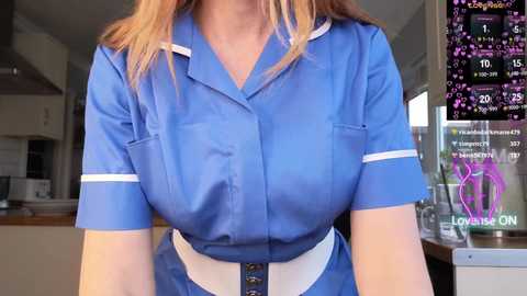 Video of a light-skinned woman with shoulder-length blonde hair wearing a blue nurse uniform with white trim, white belt, and a clipboard in a modern kitchen.