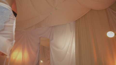 A video of a dimly lit, cozy bedroom with soft, warm lighting. The ceiling is draped with white fabric, creating a canopy effect. The bed is covered in white sheets, and a small round lamp glows softly in the background.