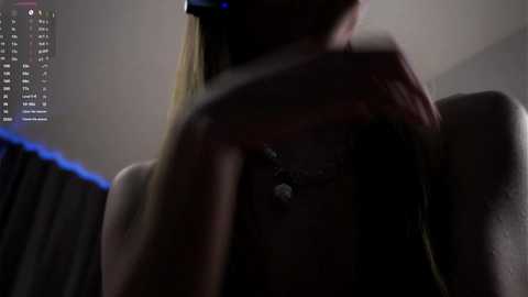 A dimly lit video of a woman with light skin and long blonde hair, wearing a black top, with a necklace and a black mask covering her face. Background shows a blurred, dark room with a computer screen displaying text.