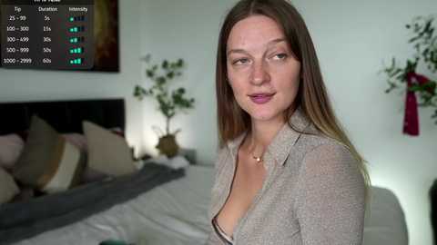 Video of a fair-skinned woman with long brown hair, wearing a gray cardigan, standing in a modern bedroom with a large bed, green potted plant, and a digital clock showing 8:07.
