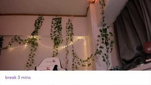 Video of a cozy, dimly-lit room with white walls adorned with green ivy and fairy lights. A white gaming chair with red accents is in the foreground, next to a table with a red apple.