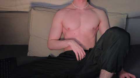 Video of a shirtless, fair-skinned man with a slim physique, wearing black pants and a pearl necklace, sitting on a bed with beige pillows and a dark bedspread.