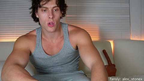Video of a muscular, light-skinned man with short, curly hair, wearing a gray tank top and jeans, sitting on a white couch with blinds in the background.