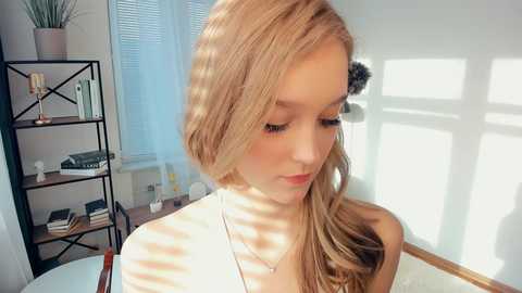 Video of a young, fair-skinned blonde woman with wavy hair, wearing a white top, standing indoors in a bright room with sunlight streaming through blinds. Background includes a black metal bookshelf and a window.