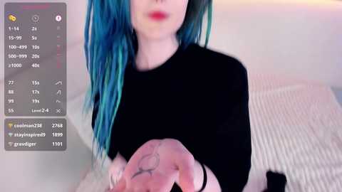 Media: Video of a fair-skinned woman with striking blue hair, wearing a black top, holding her hand with a tattoo, in a bedroom setting.