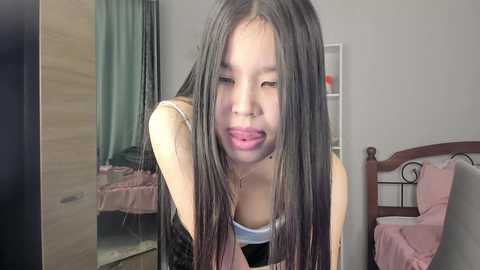 Video of an Asian woman with long black hair, wearing a white tank top, sticking her tongue out in a playful manner, standing in a cluttered bedroom with a bed and wooden furniture.