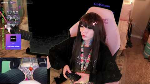 A video captures a young woman with long, straight dark hair, wearing a black hoodie, playing a video game on a pink gaming chair. She sits in a cozy, dimly-lit room.