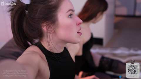 A video of a young woman with fair skin, brown hair in a ponytail, wearing a black sleeveless top, seated on a bed in a dimly lit room.