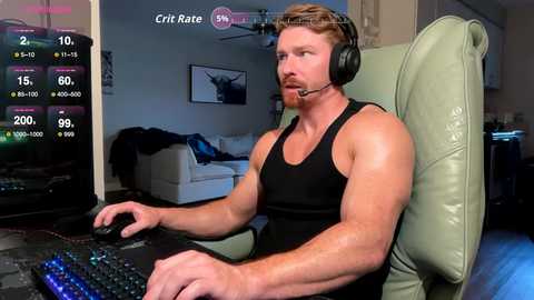 Video of a muscular, bearded man with short blonde hair, wearing a black tank top and headphones, seated at a gaming PC in a dimly lit room.
