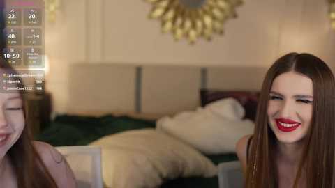 Video of two smiling women, one brunette with red lipstick, in a modern bedroom with a bed and gold wall mirror.