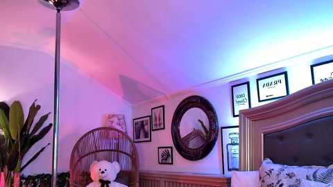 A cozy bedroom with a wicker chair, plush teddy bear, round mirror, framed art, and a bed with a tufted headboard under a soft blue and pink light.