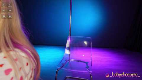 Video of a blonde woman in a red-patterned dress, standing in front of a clear plastic chair under a blue and purple stage light, with \"babychorople\" watermark in the bottom right.