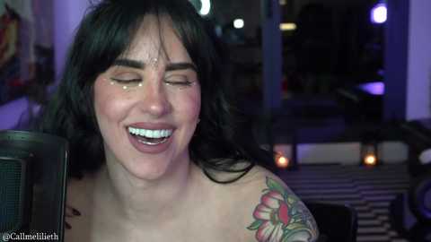 Video of a smiling woman with dark hair, freckles, and a colorful tattoo on her shoulder, wearing a microphone, in a dimly lit room with blurred background objects.