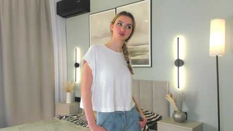 Video of a young Caucasian woman with light skin, brown hair, and glasses, wearing a white t-shirt and blue jeans, standing in a modern, minimalist bedroom with grey walls, two wall sconces, and a zebra-print bedspread.