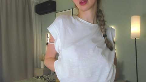 Video of a Caucasian woman with light skin, wearing a white t-shirt, braided hair, and red lipstick. She stands in a dimly lit room with a beige wall, a floor lamp, and zebra-patterned fabric in the background.