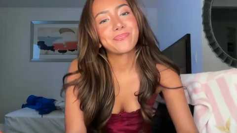 Video of a smiling, slender, light-skinned woman with long, wavy brown hair, wearing a red lace bra, sitting on a bed in a bedroom with a framed abstract painting on the wall.
