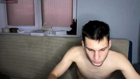 Video of a shirtless young Caucasian man with short dark hair, sitting on a beige couch, looking down, in a dimly lit room with a window and objects on the sill.