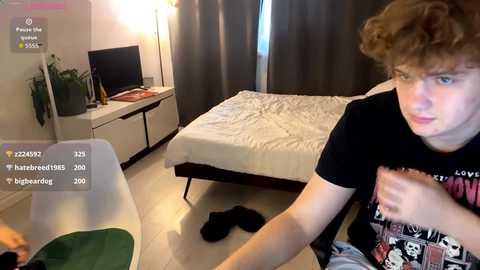 Media: A video of a young man with curly brown hair wearing a black t-shirt, sitting in a modern bedroom with a white bed, TV, and plants.