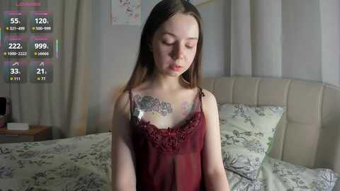 A video of a young Caucasian woman with long brown hair, wearing a sheer burgundy lace camisole, standing in a bedroom with white bedding, curtains, and a headboard.