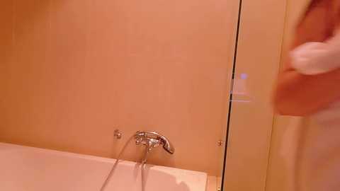 A video of a bathroom corner, showing a beige-tiled wall with a chrome showerhead and faucet, a fluffy white towel on the right, and a partially visible person.
