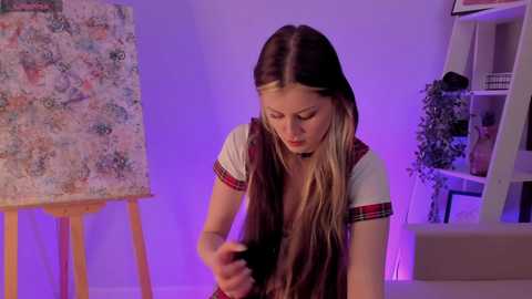 Video of a young Caucasian woman with long, straight, light brown hair styled in a side braid. She wears a white t-shirt with red plaid trim. She's painting on an easel in a softly lit room with purple lighting.