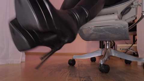 Video of a person's legs in black high-heeled boots with stiletto heels, wearing sheer black tights, standing next to a grey office chair with metal base on a wooden floor, in a room with pink walls.