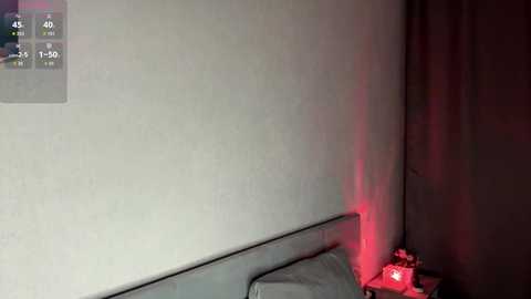 A video of a dimly lit, minimalist bedroom corner with a grey wall, a bed with grey sheets, a digital clock on the wall, and a small red light emitting from the corner, creating a mysterious ambiance.