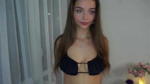 A video of a young woman with long, straight brown hair, wearing a black lace bra, standing against a white curtain backdrop. She has fair skin and a slight smile.