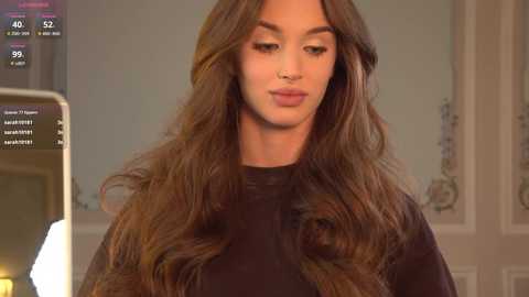 Video of a young woman with long, wavy brown hair, wearing a black robe, in a dimly lit room with floral wallpaper.