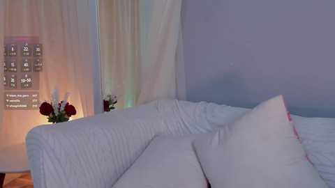 Video of a minimalist, modern bedroom with a white sectional sofa, white bedding, and a light gray wall. A decorative white and pink flower arrangement sits on a side table with a glowing, soft light.