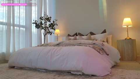 Video of a minimalist bedroom with a plush white bed, soft lighting, and a large, leafy artificial tree in the corner.