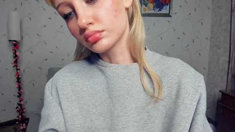 Video of a fair-skinned, blonde woman with straight hair, wearing a light gray sweatshirt, in a room with patterned wallpaper and a colorful painting on the wall.