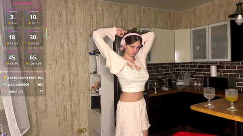 Video of a slender, light-skinned woman with long brown hair in a pink headband, adjusting her bra in a dimly lit kitchen with beige wallpaper, white cabinets, and a wooden countertop.