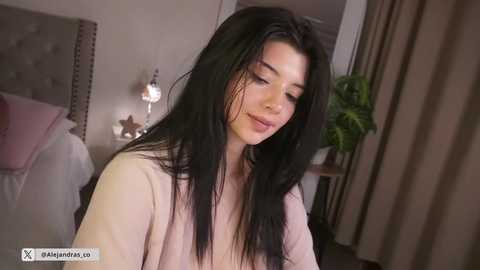 Video of a young woman with long, dark hair, wearing a light-colored robe, standing in a cozy bedroom with a bed, lamp, and green plant in the background.