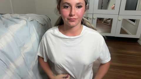 Video of a Caucasian woman with light brown hair, wearing a white T-shirt, standing in a bedroom with light-colored walls and wooden floor, next to a white wardrobe mirror.
