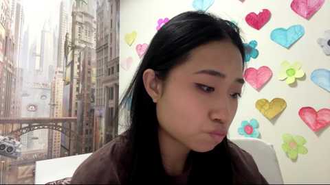 Video of a young Asian woman with long black hair, wearing a brown shirt, in a room with a mural and colorful heart cutouts on the wall.