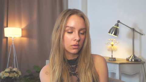 Video of a young, blonde woman with long hair, wearing a black lace top, in a modern room with beige curtains, a lit lamp, and a desk lamp.